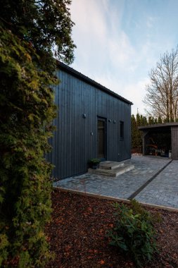 Alacakaranlıkta koyu renk kabin girişinde iki küçük lamba, bir taş yol ve alçak beton veranda görünüyor. Çıplak dalları ve yaprakları olan, budanmış çitlerin yanında bir kulübe duruyor..