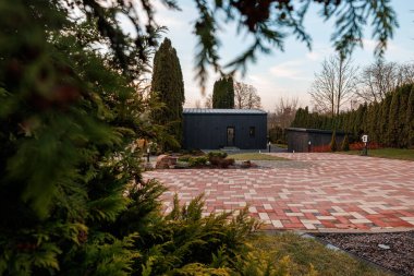 Metal çatılı Matte Black Stüdyosu ringa kemiği geçitlerinin, kozalakların ve çitlerin ötesinde yer alır. Beyaz bir EV şarj aleti çimlerin yanında duruyor. Gün ışığı yumuşak gölgeler bırakıyor..