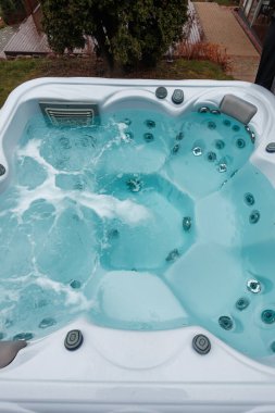 Verandadaki beyaz dikdörtgen jakuzinin yukarıdan görünüşü. Turkuaz su kabarcıkları, krom jetler ve kontroller kenar çizgisinde. Bulutlu ışık serinlik katar.
