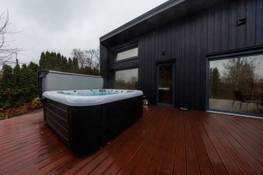 Dikdörtgen şeklinde bir jakuzi, koyu renk bir evin yanındaki, kırmızı renkli kahverengi güvertede buharlaşır. Sürgülü cam kapılar yapraksız ağaçları yansıtır. Serin sonbahar ışığı ve geniş açılı çerçeve..