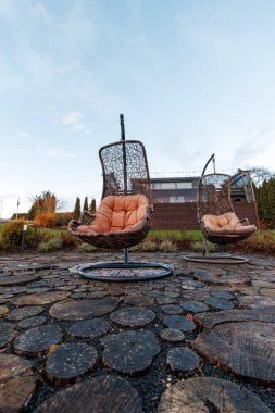 Bronz yastıklı iki hasır yumurta koltuğu, ahşap mermilerden oluşan bir avluda asılı duruyor, düşük açılı serin gün ışığı, desenli ahşap ve örülmüş kafes, yakındaki tel lambaları..