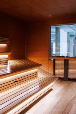 Sedir banklı boş sauna, LED şeritler, ve büyük cam bölmeli metal soba kapalı bir spa, geniş açılı dikey çekim ve sıcak ışıklandırma..