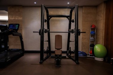 Smith makinesi, bank, koşu bandı ve eğitim teçhizatı ile sıkışık spor salonu. Yumuşak teneffüs aydınlatma ve taş dokulu duvarlar, düzenli çalışma saatlerinde cilalı bir titreşim oluşturur..