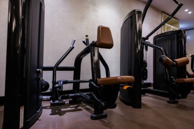 Kahverengi pedli bir dizi siyah seçilmiş makine modern bir spor salonunda duruyor. Yastıklı koltuklar ve sarı iğneli ağırlık yığınları yumuşak, hatta ışık altında bile görülebilir..
