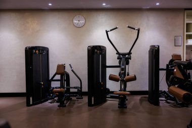 Modern jimnastik salonu oturma sırasını, çift kollu omuz baskısını ve seçilmiş birimleri gösteriyor. Beyaz bir analog saat 10 'u gösteriyor. Aralıklı ışıklar parlayan zemini aydınlatıyor..