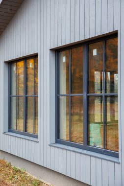 Uzun pencereli ve gri kaplamalı modern ev cephesi. Yumuşak bulutlu ışık, açılı manzara, ılık sonbahar yansımaları, iç kutular yenilemeyi öneriyor.