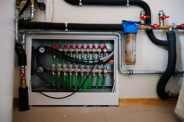 Ölçüler, kırmızı valfler ve yeşil PEX çizgileri olan duvar monte edilmiş manifoldlar, artı mavi tüp filtre ve yalıtılmış borular, kapalı alanda hidronik ısıtma ve su arıtma olduğunu gösteriyor..