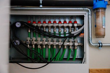 Durgun bir dolapta kırmızı akış sayaçları, göstergeleri ve yeşil PEX döngüleri olan paslanmaz bir manifold bulunur. Galvanize borular sağdaki mavi tepeli tortu filtresine bağlanır.