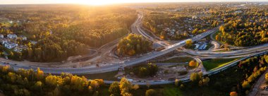 Hava panorama, öğleden sonra ilerleyen saatlerde, süpürme rampaları, yüksek bir üst geçit, kazılı koridorlar, sonbahar ormanları ve banliyö konutları ile inşaat halindeki bir kavşağı gösteriyor..