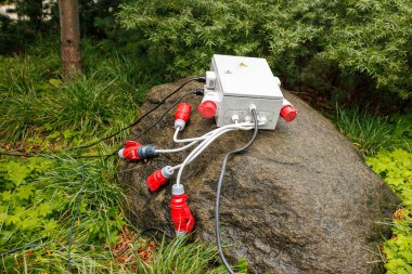 Bahçedeki bir kayanın üzerinde kırmızı ve gri CEE bağlantıları olan taşınabilir bir elektrik kutusu duruyor. Bir uyarı üçgeni yüksek voltaj gösteriyor. Islak yüzeyler yakın zamanda yağdığını gösteriyor..