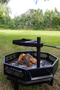 Kafes kesikli siyah bir octagonal ocak, çitlerle çevrili bir çimenlikte, bir kazık ve ahşap saplı döner ızgara ile bölünmüş odun ve kül tutar..