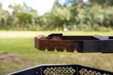 Paslı ateş çukuru, birbirine kenetlenmiş çentik ve eğri şeritli başlık. Yakından bakınca altıgen duvarın üzerinde çelik çember görünüyor. Kafes kesikleri, yumuşak gün ışığı, sığ derinlik, yıpranmış gübre..