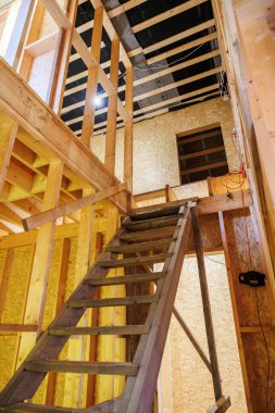 Tamamlanmamış ahşap çerçeve ve dik geçici merdiven. Açık çiviler, OSB kılıfları, elektrik tesisatı, iş lambası ve siyah buhar bariyeri olan kirişler aktif inşaatı gösteriyor..