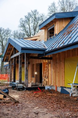 Kısmen inşa edilmiş ahşap kulübe mavi ayakta dikişli metal çatı, çivili çiviler, derin saçaklar ve çamurlu bir sitede merdivenler, aletler ve parlayan bir kapı.