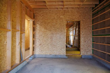 OSB duvarları ve tavanı, yeşil bant, açıkta duran çiviler, kirişler, kapı aralıkları ve çiğ beton kaplamayla inşa edilmiş bir Kuzey Amerika evinin gündüz görüntüsü..