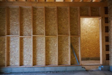 Tamamlanmamış bir iç mekan dikey 2x4 çiviler, OSB kılıfları ve beton blok bir kök duvarda pürüzlü bir kapı gösteriyor. Ilık tungsten ışığı yayılıyor, yaslanan bir kanal borusunu vurguluyor..