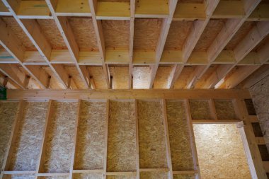 Taze çivili duvarlar ve OSB kılıflı boyut kirişleri inşaat ışıklarıyla aydınlatılıyor. Sağda açık bir kapı var ve yük bakımını engelliyor..