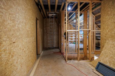 Kısmen inşa edilmiş tavan arası çivileri, OSB duvarları ve asılı iş lambaları olan kirişleri gösteriyor. Metal merdiven, giriş kapağıyla birleşir. Kablolar ve aletler alt katta yatar..