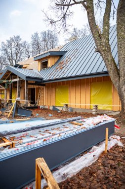 Çağdaş ahşap ev yaprak döşeme, gri ayakta dikiş çatısı, sarı plastik pencereler, gündüz yaprak sahasında merdivenler ve testere atlarıyla inşa ediliyor..