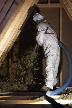 Beyaz takım elbiseli bir montajcı inşaat sırasında bitmemiş bir tavan arasına selüloz üflemek için mavi bir hortum kullanır..