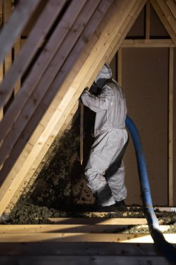 Beyaz elbiseli bir işçi mavi bir hortumdan selüloz yalıtımını üfleyerek yeni ahşap çerçeveli ve havada gevşek lifler bulunan dik üçgen tavan arasına fırlatır..