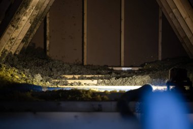 Bir müteahhit tavan arasındaki kirişler ve koyu renk kılıflar ve malzeme hortumları ile kirişler arasında patlayan selüloz yalıtımı aydınlatıyor..