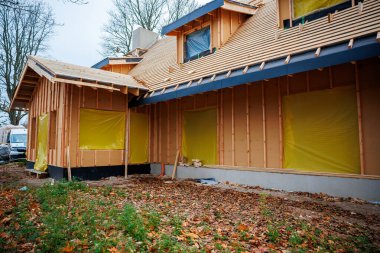 Kısmen inşa edilmiş konutlarda çiviler, kılıflar ve yarasalar görülüyor. Sarı film pencereleri kapatıyor, tepenin yanında baca yükseliyor, minibüs park halinde, sonbahar ağaçları ve yapraklar var..