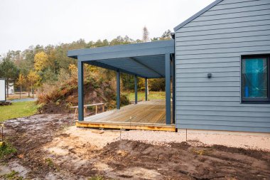 Gri kaplamalı ve çelik mavi pergola güverteli modern bir ev, sonbahar sonbaharının sonlarında ıslak tahtalar ve dağınık ışık, arka planda görünen devam eden yapı..