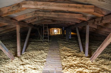 Tavan arası gibi bir tünel sert testereli kirişler, çapraz teller ve gevşek dolgu yalıtımı üzerinde dar bir tahta iskelet gösterir. Aydınlatılmış bir kapak, kablolar ve küçük bir geçit görünüyor..