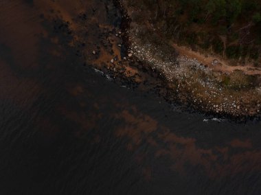 Hava görüntüsü koyu çay renkli su, demir zengini girdaplar, ve dar bir yuvarlak kaya ve çakıl kuşağı gösteriyor, çam ve çalılarla, soluk patika, serin yayılmış ışık.