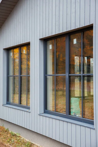Uzun pencereli ve gri kaplamalı modern ev cephesi. Yumuşak bulutlu ışık, açılı manzara, ılık sonbahar yansımaları, iç kutular yenilemeyi öneriyor.