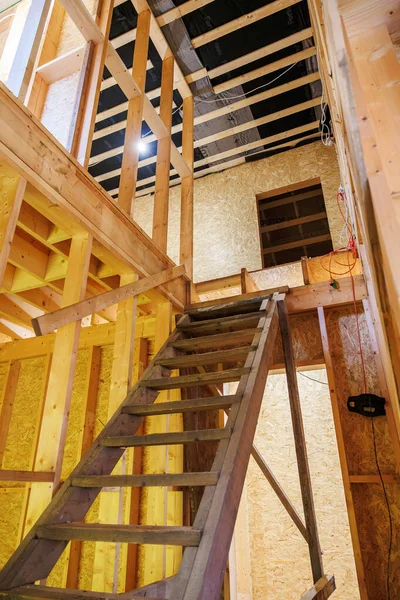 Tamamlanmamış ahşap çerçeve ve dik geçici merdiven. Açık çiviler, OSB kılıfları, elektrik tesisatı, iş lambası ve siyah buhar bariyeri olan kirişler aktif inşaatı gösteriyor..