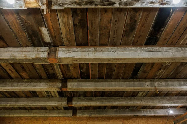 Elle yontulmuş kirişler ve karartılmış kalaslarla eski ahşap bir tavanı yakından görmek, testere izleri, lekeler ve çürüme kenarlarında küçük LED ampuller, sıcak düşük ışık, yukarı çerçeve.