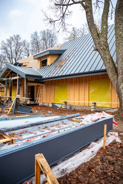 Çağdaş ahşap ev yaprak döşeme, gri ayakta dikiş çatısı, sarı plastik pencereler, gündüz yaprak sahasında merdivenler ve testere atlarıyla inşa ediliyor..