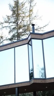 Ahşap kaplı evin üst köşesinde ayna kaplaması ve bacası olan siyah çelik sütunlar ve aşağı ışıklar. Karaçam ağaçları, rüzgarın öğleden sonra hareket ettiği gibi yansıyor..