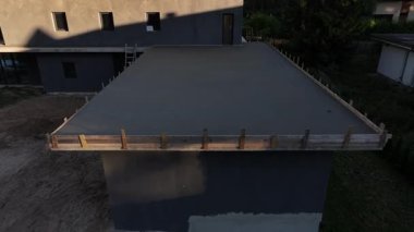 Yeni banliyö konut inşaatında havadan beton bir çatı levhası. Kereste işleri, merdiven, dikdörtgen pencereler, tahta çitler, olgun kozalaklı ağaçlar. Yavaş dron hareketi, drenaj çıkışı, merdiven, uzun gölge..