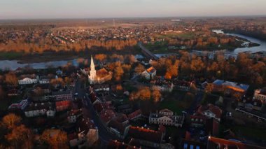 Beyaz kilise havası, yüksek kule ve kıvrımlı nehir başlığı. Kırmızı kiremit çatılar dar sokaklar arasında toplanır, taş köprü öğleden sonra aydınlığında çöker..