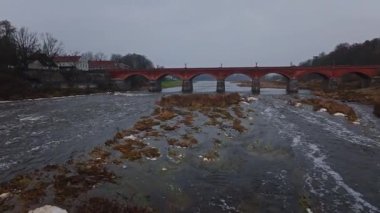 Kışın Venta Nehri üzerindeki Kuldiga Havalimanı kırmızı tuğla kemer köprüsü. Yavaş hareket taş iskeleleri, kamış adalarını, küçük akıntıları, Letonya renklerine sahip sokak lambalarını ve eski şehir çatılarını gösterir..