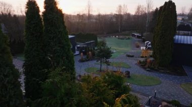 Hava görüntüsü kompakt avlu, modern küp ek binalar, desenli tuğla geçitler, havuz, oyun alanı, huş ağaçları, pergola sicim ışıkları ve kamera dışa dönük.