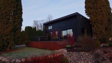 Hava aracı tavası, eğimli çatılı ve yükseltilmiş güverteli modern siyah giysili bir ev gösteriyor. Kırmızı dal ışıkları ve ampuller parıldıyor, jakuzi ve sandalyeler dışarıda oturuyor, iç ışık gösteriyor..