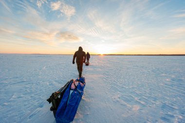 İki balıkçı gün doğumunda donmuş bir alanda yürürler, süspansuvarlarla kızakları çekerler, uçlarını yukarı çekerler ve muşamba kaplı bir takım. Uzun gölgeler ve uzak bir ağaçlık çerçeve Geniş bir manzara.