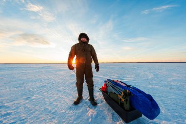 Bir bohça balıkçı, güneş doğarken geniş donmuş bir gölde siyah bir kızak çeker. Uzun turuncu göller sabah erken saatlerde dalgalanan karları işaret eder..