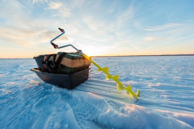 Sarı bıçaklı bir el kumandası güneş doğarken geniş donmuş bir gölde siyah bir dişli kızağın yanında durur. Taze kızak izleri bir balıkçıyı işaret ediyor. Kıtır kar üzerinde ılık ışık parıldıyor.