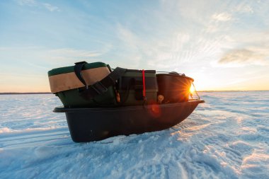 Güneş doğarken donuk bir gölün üzerinde, yeşil takım çantalı, kovalı ve kayışlı bir vites kızağının düşük açılı görüntüsü, uzun gölgeler soğuk görünüyor.