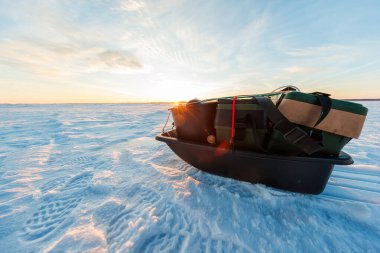 Düşük açı, buz parçalarının üzerinde alet çantaları, kova ve kayışlar olan siyah bir kızak gösteriyor. Çizme izleri ve çekme ipi kışın altın saatinde duraksadığını gösteriyor..