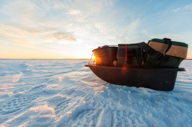 Düşük açılı, takım çantalı, kovalı siyah bir kızak ve güneş doğarken karla kaplı göl buzuna bağlanmış kayışlar. Lens ve uzun gölgeler kışın ortasında olduğunu gösteriyor..