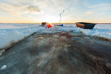 Islak çamur ve kırmızımsı yem lekeleriyle taze bir buz deliği gün batımında uzun ve sıcak bir ışıkla birlikte geniş donmuş bir göle el çapası, kova, kızak ve dişli taşır..