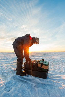 Donmuş bir gölün üzerinde gün doğumunda bir dişli kızağın üzerine eğilir, ceketin içinde sıcak bir ışık parlar, uzak bir ağaç hattının yanında uzun ışınlar uzanır..