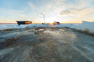 Düşük açılı kış sahnesinde yeni delinmiş bir göl deliği, yeşil bir el çapası, bir kızak ve teçhizat gün doğumunda arka planda, çıkıntılı kar kütleleri ve ıslak buzlar görülebilir..