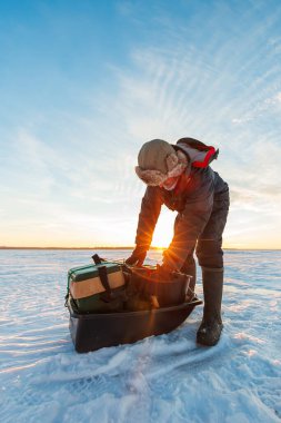 Kürk şapka ve bot sıralama bir bohça balıkçı çubuklar, çapa, ve donmuş bir göl üzerinde siyah bir kızak mücadele gün doğumunda, güneş kol ve buz ile parlıyor.